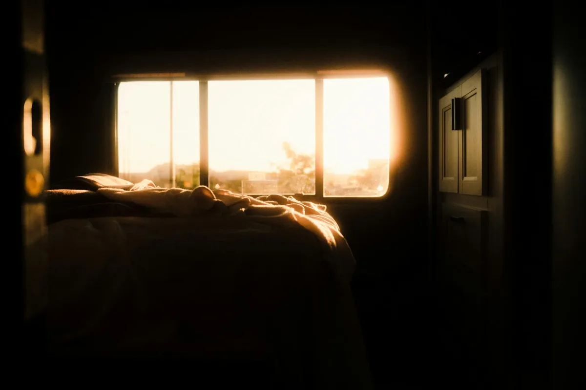 A bedroom at dawn, sheets rumpled, window closed, a thin line of light through curtains.