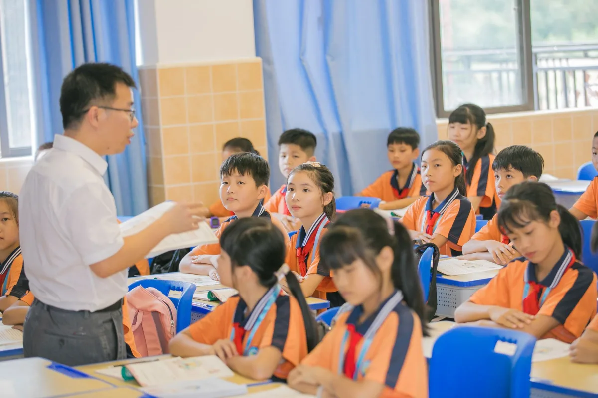 An elementary school classroom with desks, daylight from large windows, and a chalkboard.