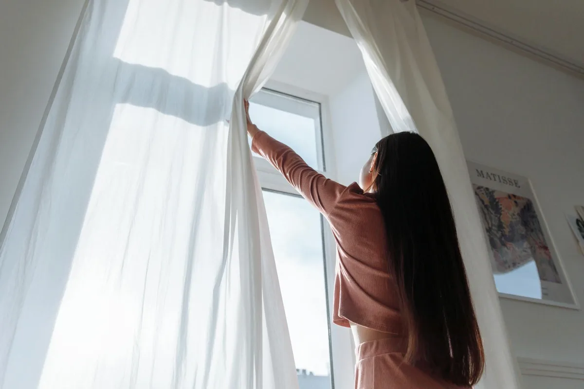 A casement window cracked open onto a sunny morning, sheer curtain fluttering inward.
