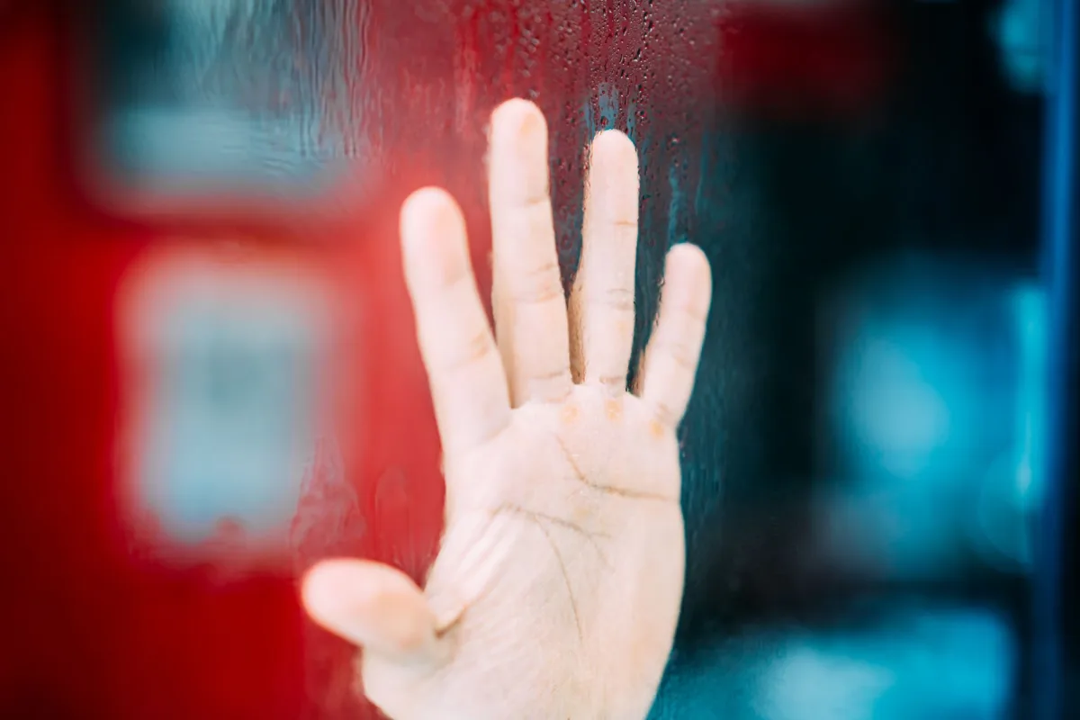 A close-up of someone with hands on a windowpane, looking thoughtful.