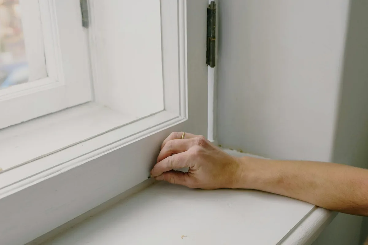 A gloved hand wiping a thin layer of pale ash off a windowsill with a damp microfiber cloth, with a HEPA vacuum on the floor behind.
